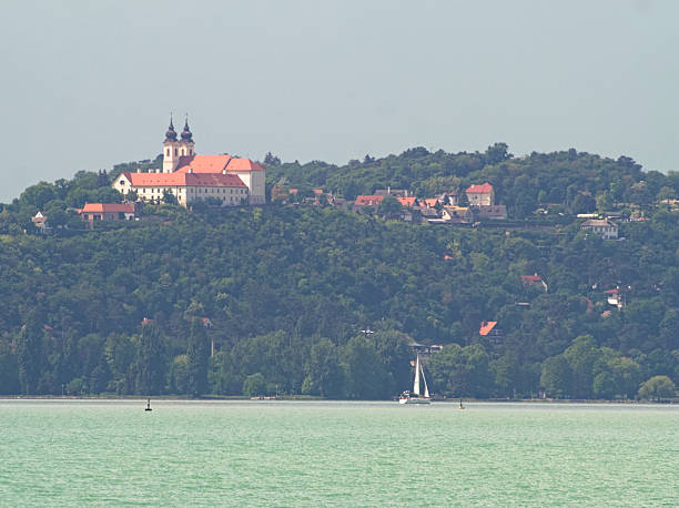 A scenic panoramic view of the Tihany Peninsula on Lake Balaton, showcasing the historic Tihany Abbey atop a tree-covered hill, with a white sailboat gracefully gliding on the lake's calm waters.