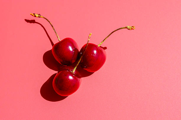 Three glistening red cherries with long stems lie on a bright pink background, creating contrast and interesting shadows. This minimalistic composition is ideal for design, advertising, or as a backdrop for culinary themes