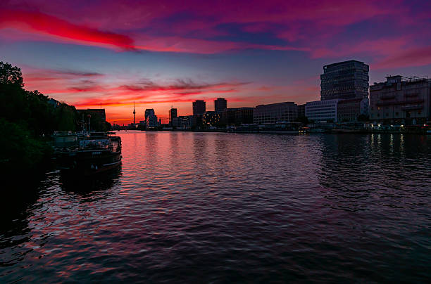 Editorial stock photo taken from Oberbaum Bridge in Berlin, showing the Spree River at dusk. The vibrant sky displays intense red, pink, and purple tones as night begins to fall. A single boat is anchored on the left riverside, while city lights start to reflect on the calm water. Berlin’s skyline with the iconic TV Tower and modern architecture completes the tranquil urban scene.