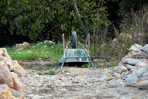 A rusted, overturned wheelbarrow with faded paint lies unused on rocky, uneven ground, surrounded by wild grass and stones, evoking a sense of forgotten rural labor