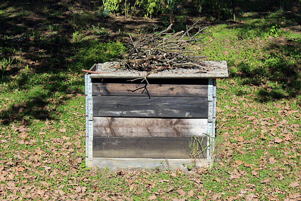 A square, homemade compost bin built from weathered wood and metal brackets sits on a leafy lawn, topped with a pile of dry sticks and surrounded by green grass