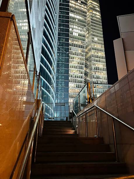 Street stairs leading towards towering blue yellow and white skyscraper buildings with bright glowing windows and reflections on the walls, shining light at night