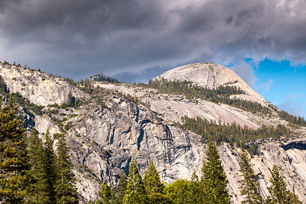 A scenic view of a mountain range with a mix of rocky terrain and lush green trees, set against a backdrop of dramatic, cloudy skies.