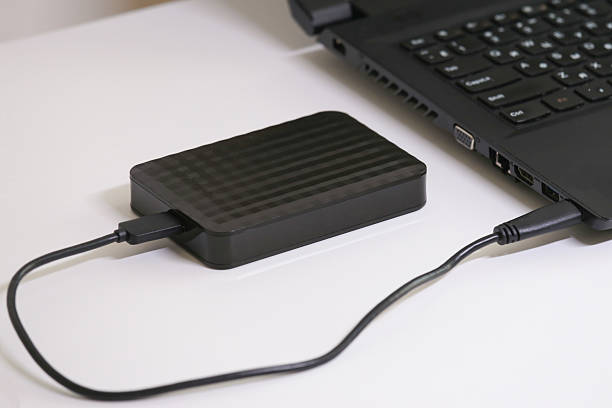 istock External hard drive connected to a laptop via USB cable on a white surface. Portable data storage device for backup, file transfer, and secure digital information management. 2223760283