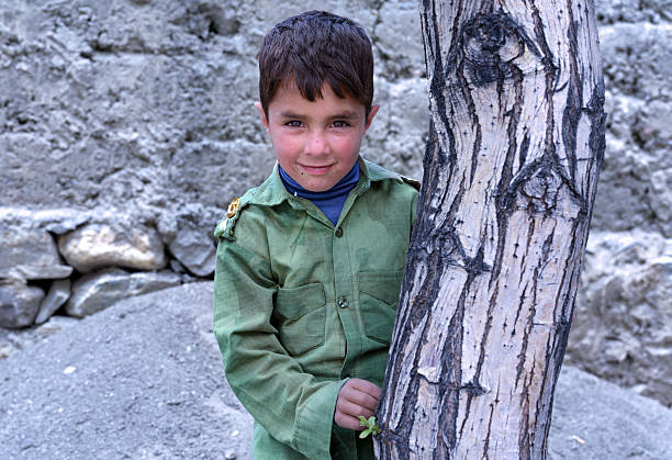 Portrait of a smiling afghan boy wearing a military-style jacket, standing next to a tree in a rural village