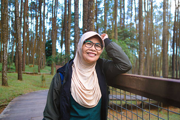 A cheerful woman wearing glasses and a hijab enjoys a serene walk through a forest of towering pine trees, exuding joy and relaxation in the tranquil natural environment