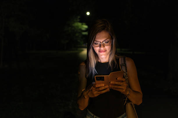 istock Beautiful blond woman walking through public park at night while her face is illuminated by screen of her mobile phone. She is browsing social networks or internet for information. 2223770773