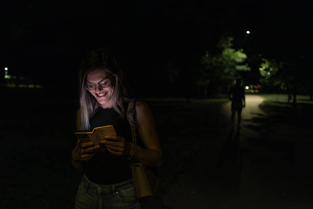 istock Beautiful blond woman walking through public park at night while her face is illuminated by screen of her mobile phone. She is browsing social networks or internet for information. 2223772356