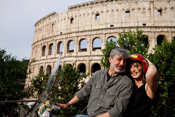 A mature couple is getting ready for a motorcycle ride in Rome, Italy. The man sits on a scooter while the woman holds a red helmet, preparing for a scenic adventure near the iconic Colosseum. A perfect moment showcasing senior travel, freedom, and tourism in Italy.