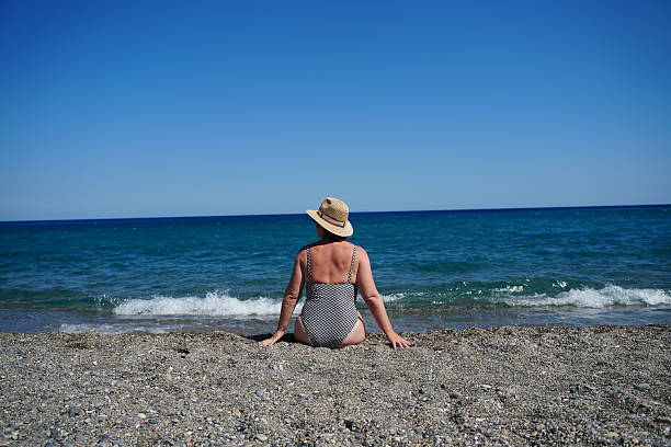 Woman sits on a pebble beach facing the ocean, wearing a hat and patterned swimsuit. Relaxing moment with waves in the background and clear blue sky above