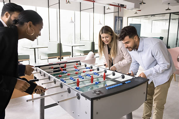 Casual office fun. Joyful enthusiastic multiethnic workmates colleagues engaged in friendly competition play foosball match divided into teams enjoy funny teambuilding activity playful spirit unity