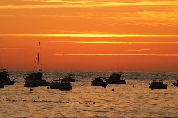 Silhouetted boats float on a tranquil sea at sunset, creating a serene and picturesque scene with vibrant orange hues in the sky.