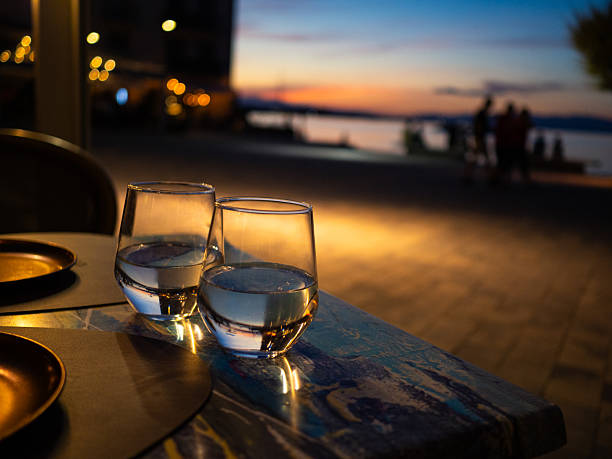 Two glasses of water sit on a table illuminated by soft evening light, with people socializing in the background against a beautiful sunset over the water.