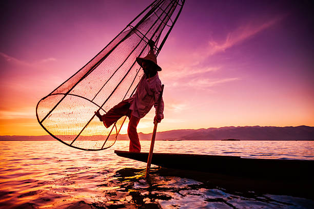 Asian fisherman on wooden boat casting a net for catching freshwater fish in nature river in the early morning before sunrise