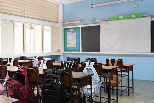 Interior of a school classroom with wooden desks, student backpacks hanging on chairs, blackboard and whiteboard at front, windows letting in bright natural sunlight, education and learning concept.