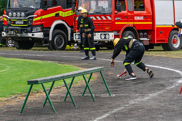 Sieroszewice, Greater Poland Voivodeship, Poland - june 29, 2025 volunteer firefighter falls while handing off the baton during a fire sport relay race. Fire trucks, teammates in helmets, and obstacle course elements are visible in the background.