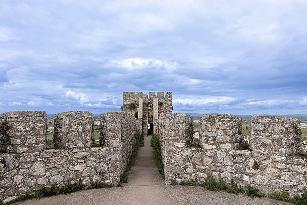 Pathway between ancient stone walls leading to a watchtower, with a cloudy sky creating a dramatic backdrop