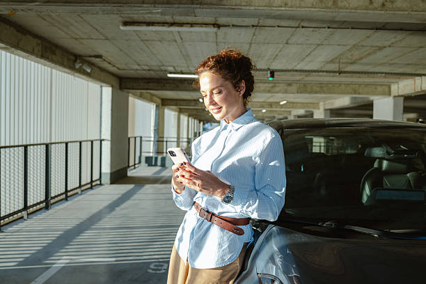 istock Smiling woman using smartphone while leaning against car in modern parking garage 2223813064