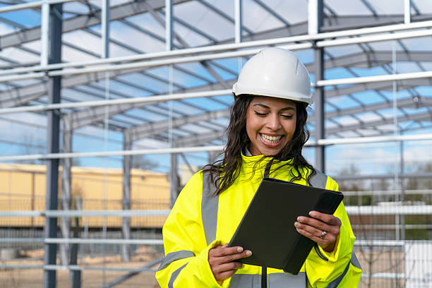 istock Portrait of happy smiling female construction worker builder with tablet, dressed in high visibility gear and helmet, standing in front of partially completed steel frame structure 2223813965