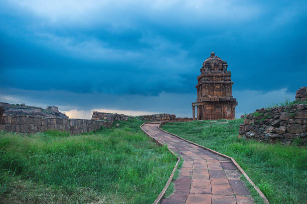 A serene walkway leads to a small ancient temple standing amid lush green grass under a dramatic cloudy sky at Badami, Karntaka, India.