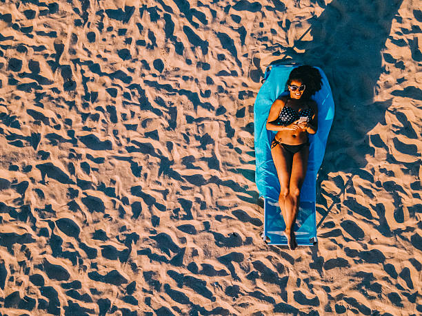 Aerial view of a smiling woman in a black bikini sunbathing on a beach lounger, surrounded by patterned sand and casting a long shadow. The vibrant scene captures the essence of summer relaxation and beach lifestyle.