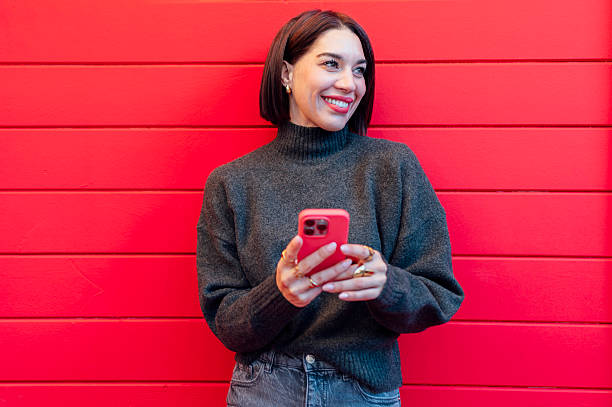 istock Young woman using smartphone and smiling, leaning against a red wall 2223823012