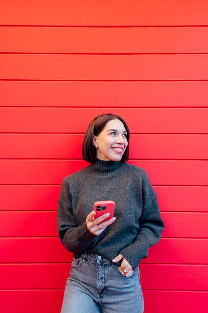 istock Young woman using mobile phone and smiling looking away while standing against red wall 2223823013