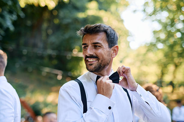 Happy groom adjusting his bow tie, smiling brightly while glancing away, preparing for the wedding ceremony in a picturesque park, surrounded by lush greenery and summer sunlight