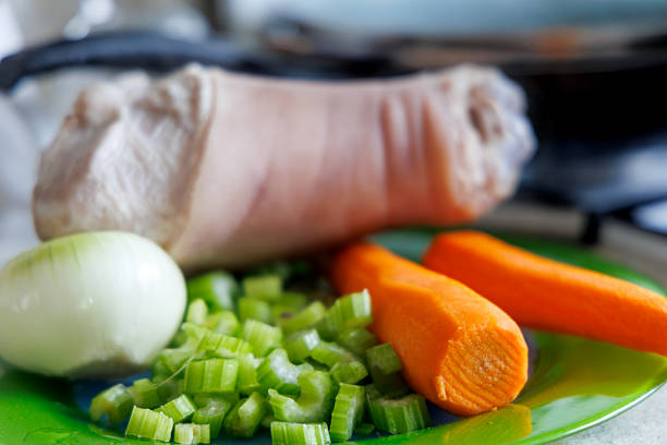 A peeled pork shank is visible on a green plate, next to it is half an onion, chopped celery stalks, and two carrots.