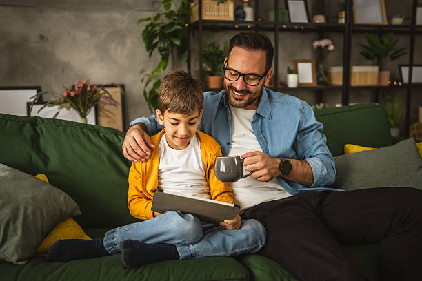 istock Father hold cup of coffee and look son while he use tablet for fun 2223849620