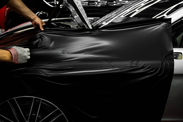 istock Car wrapping process using black matte vinyl stretched into place. Careful application and tensioning of black vinyl avoids surface damage or bubbling. Matte black vinyl is applied across car hood. 2223853035