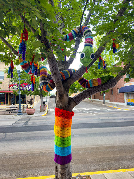 Klamath Falls Oregon/USA - June 27th 2025: This image shows a colorful knitting bomb crafted in the shape of a snake, artfully wrapped around a real tree trunk in downtown Klamath Falls, Oregon. The yarn installation, part of a vibrant urban art movement, contrasts with the natural bark texture of the tree and the surrounding urban environment. This example of yarn bombing highlights community creativity and brings unexpected color and whimsy to the city streets, blending handcrafted fiber art with public green spaces.