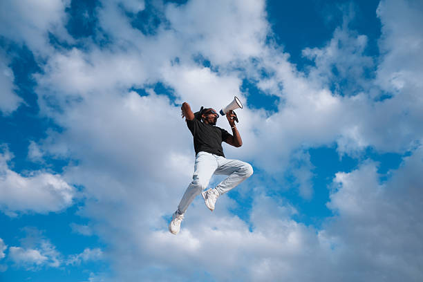A man is jumping in the air with his arms outstretched. The sky is blue and there are clouds in the background. Scene is joyful and energetic