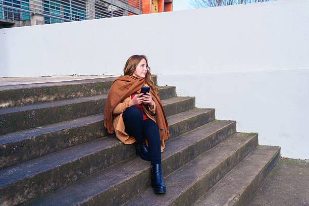 Young woman using smartphone while sitting on concrete steps in the city during winter