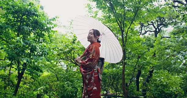 A woman in a traditional Japanese costume and carrying an umbrella walks in the garden on a rainy summer day. She smiles at the camera.