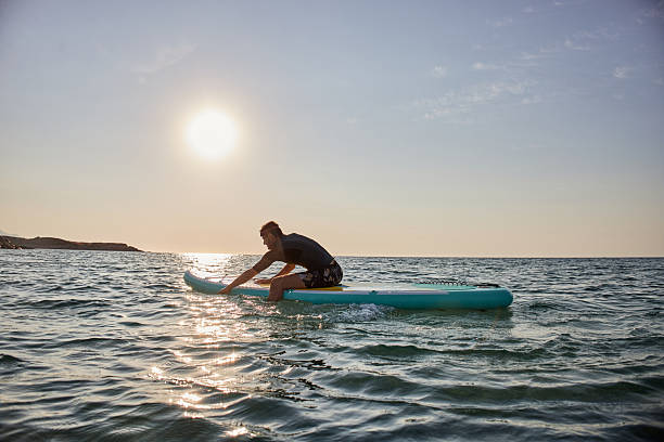 Happy young adult male surfer in a wetsuit lying on the surfboard and paddling with his hands. He is trying to catch a wave during a beautiful sunrise.
