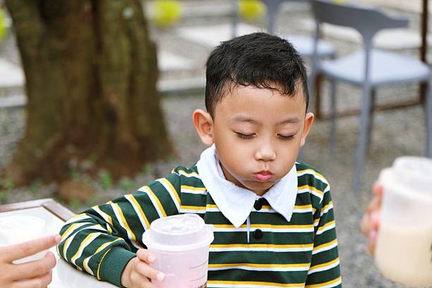 A young Asian boy in a striped shirt holds a drink while sitting outdoors. He looks calm and pensive, surrounded by natural elements and casual seating.