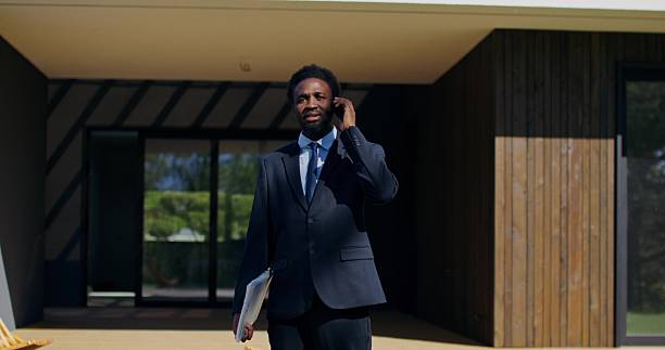 A real estate agent is talking on a mobile phone while standing near house for sale on a summer day