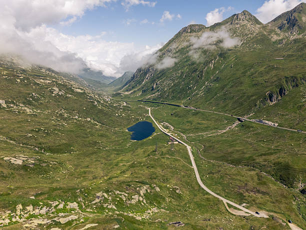 Aerial view of a winding road through a green mountain valley with a small blue lake nearby. Scenic alpine landscape with rocky hills and clouds. Mountain scenery with open nature and road.