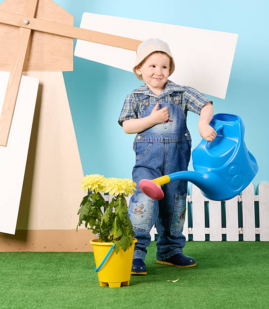 Cute red-haired boy in farm clothes standing and watering a flower from a watering can. Studio shot, blue background, green lawn, big toy mill