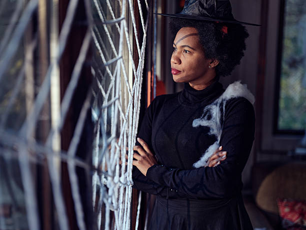 Pensive African American woman dressed up as a witch on Halloween looking through the window at home. Photographed in medium format.