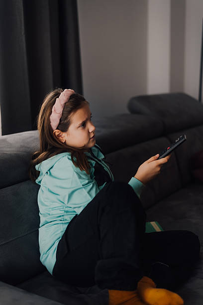 istock Young girl sitting on sofa watching television changing channels with remote control 2223886309