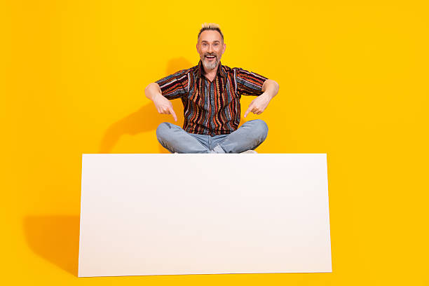 istock Charismatic man displays cheerful attitude while sitting cross legged on a colorful background with a blank sign for advertising. 2223889181