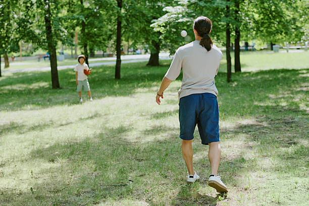 Father playing catch with young child in green park, man throwing baseball toward child holding glove, both enjoying outdoor activity surrounded by trees and grass