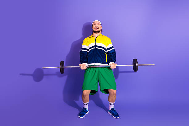 istock Young man lifting a barbell with funny expression against a solid purple background wearing retro sportswear and gym outfit 2223890212