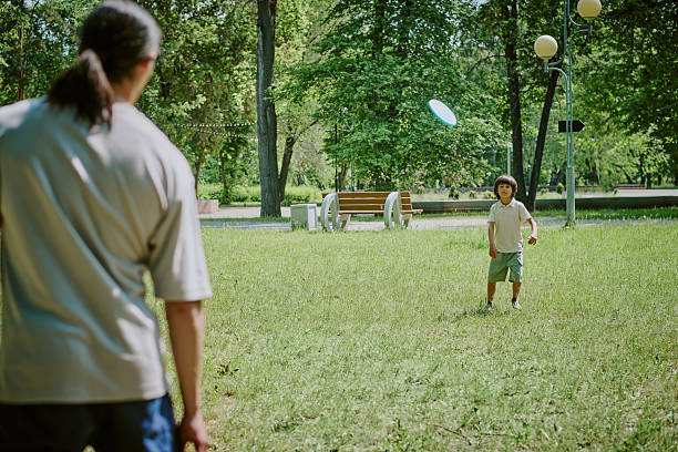 Father throwing flying disc to child boy in park, child standing on grass with arms ready to catch, trees and benches visible in background