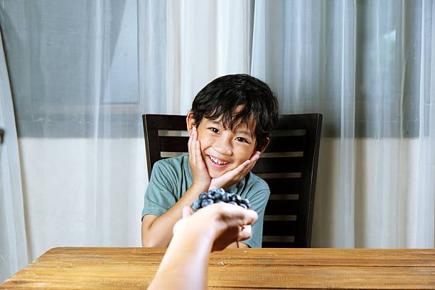 A cheerful Asian boy smiles brightly as he receives a handful of fresh blueberries, representing healthy snacking and positive food habits in childhood.