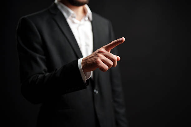 Man in suit gestures with finger pointing while standing against dark background expressing authority and confidence