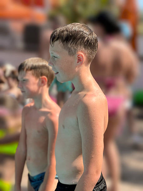 A small group of school aged children stand together pool-side as they pose together for a portrait after a swimming class.  They are each wearing swim suits and are smiling.