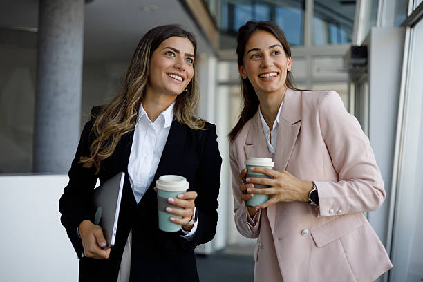 istock Professional women enjoying coffee break, discussing project details while holding laptop and sustainable mugs in corporate hallway 2223913786